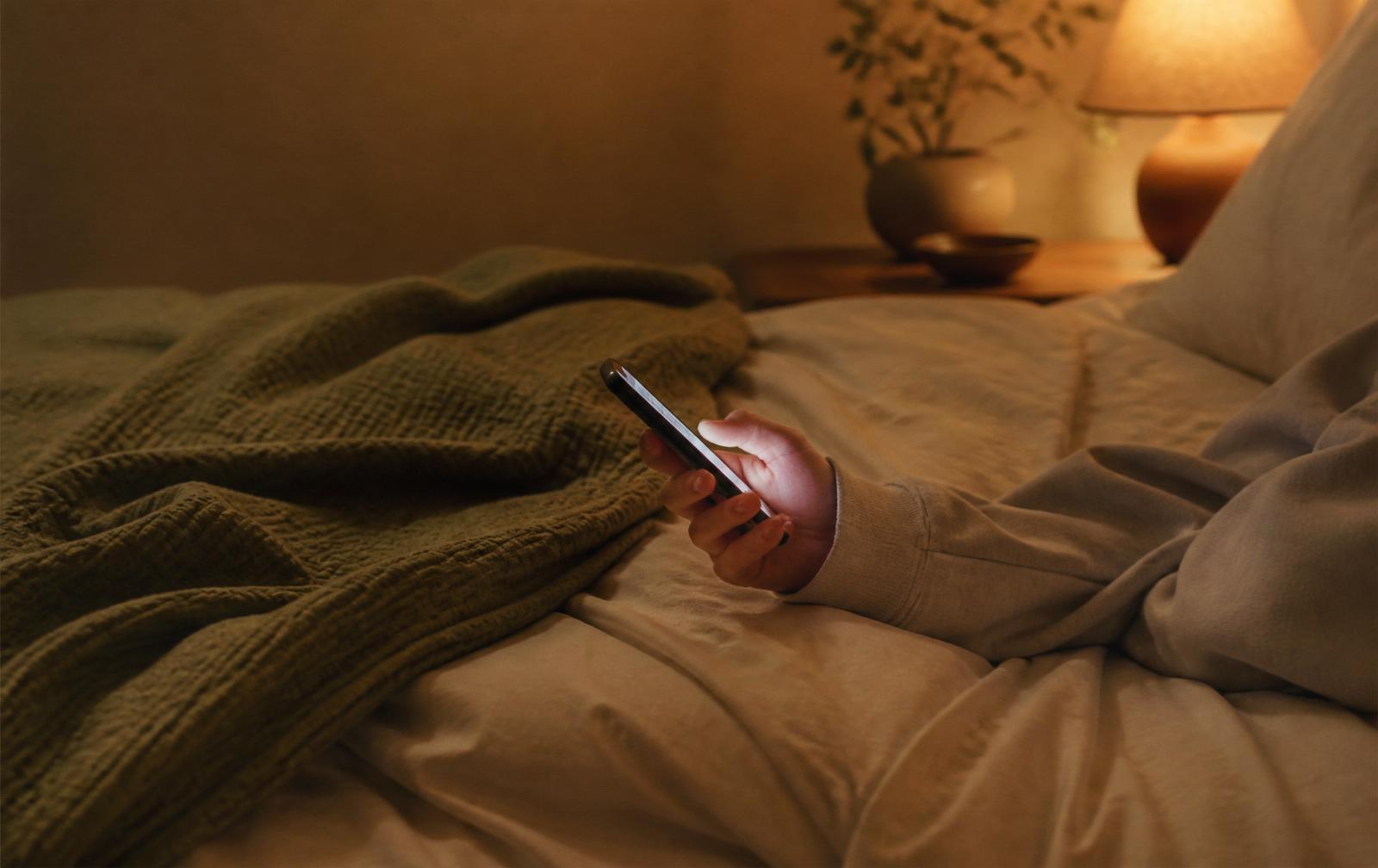 A teenager's hand holding a softly glowing phone in a warm, dimly lit bedroom with cream bedding and a sage throw